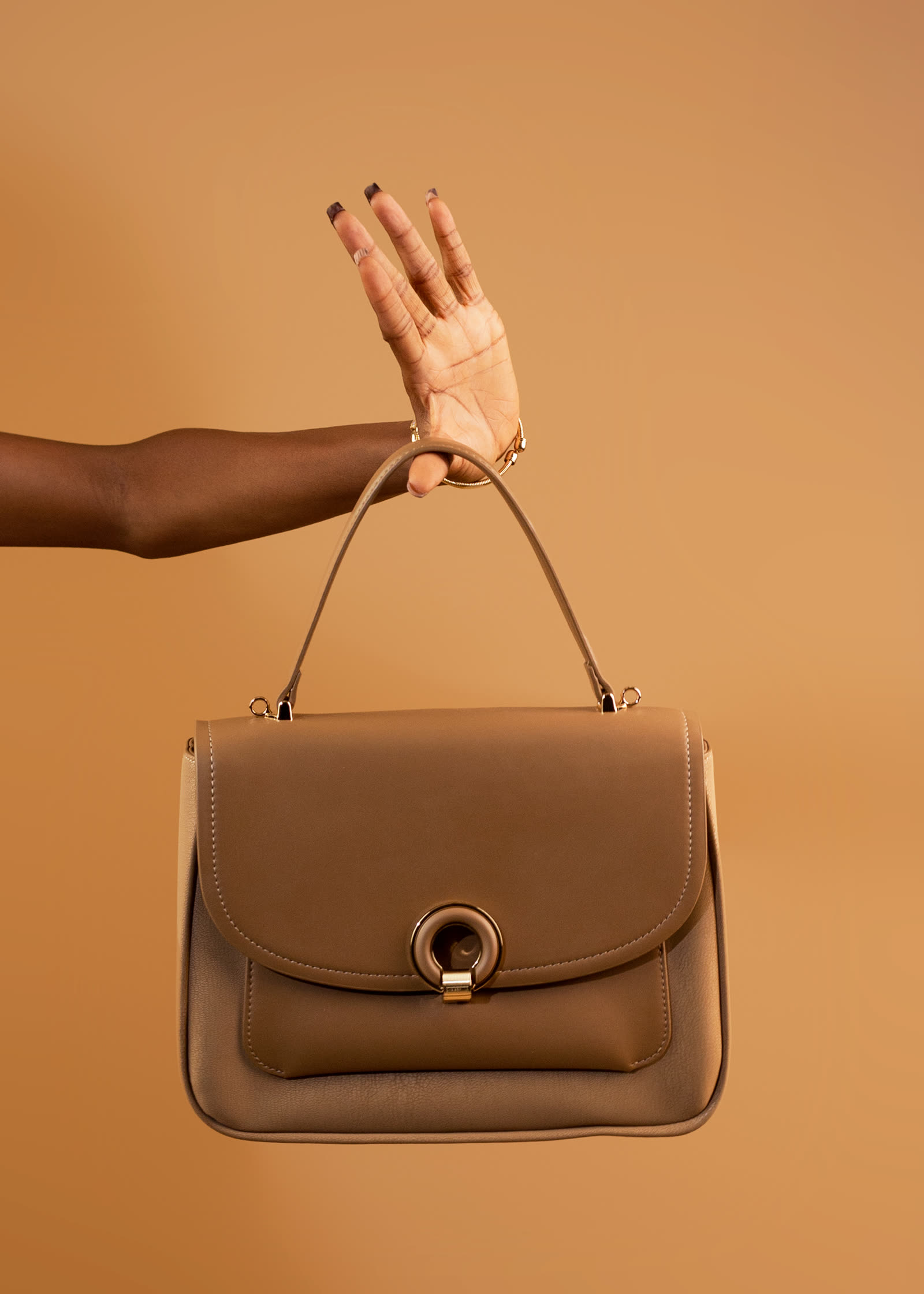 An outstretched arm holds a taupe top-handle bag with a gold ring clasp against a warm caramel backdrop.