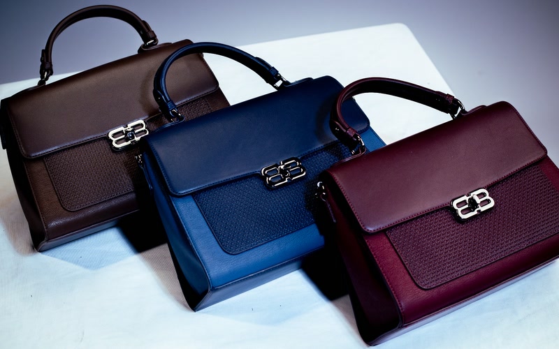 Three Bibi BX flap satchels in chocolate, navy, and burgundy, each with a woven-textured panel and gold clasp.