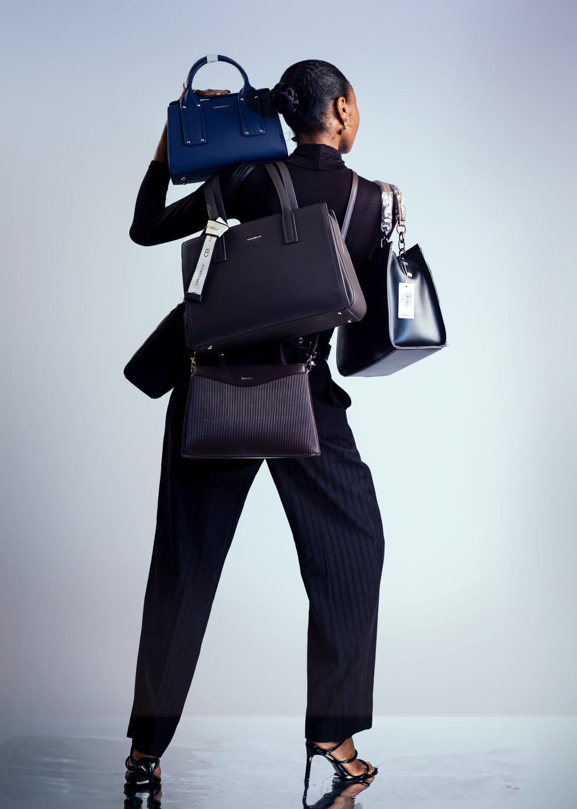 A model dressed in black carries five handbags against a pale grey studio backdrop.
